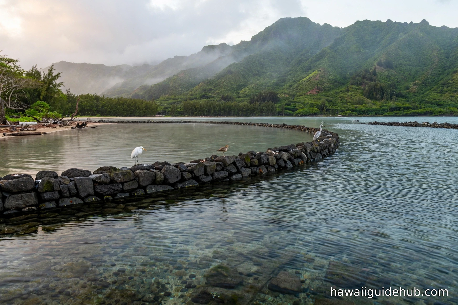 Huilua Fishpond's ancient rock wall curving through Kahana Bay with misty Koʻolau mountains behind