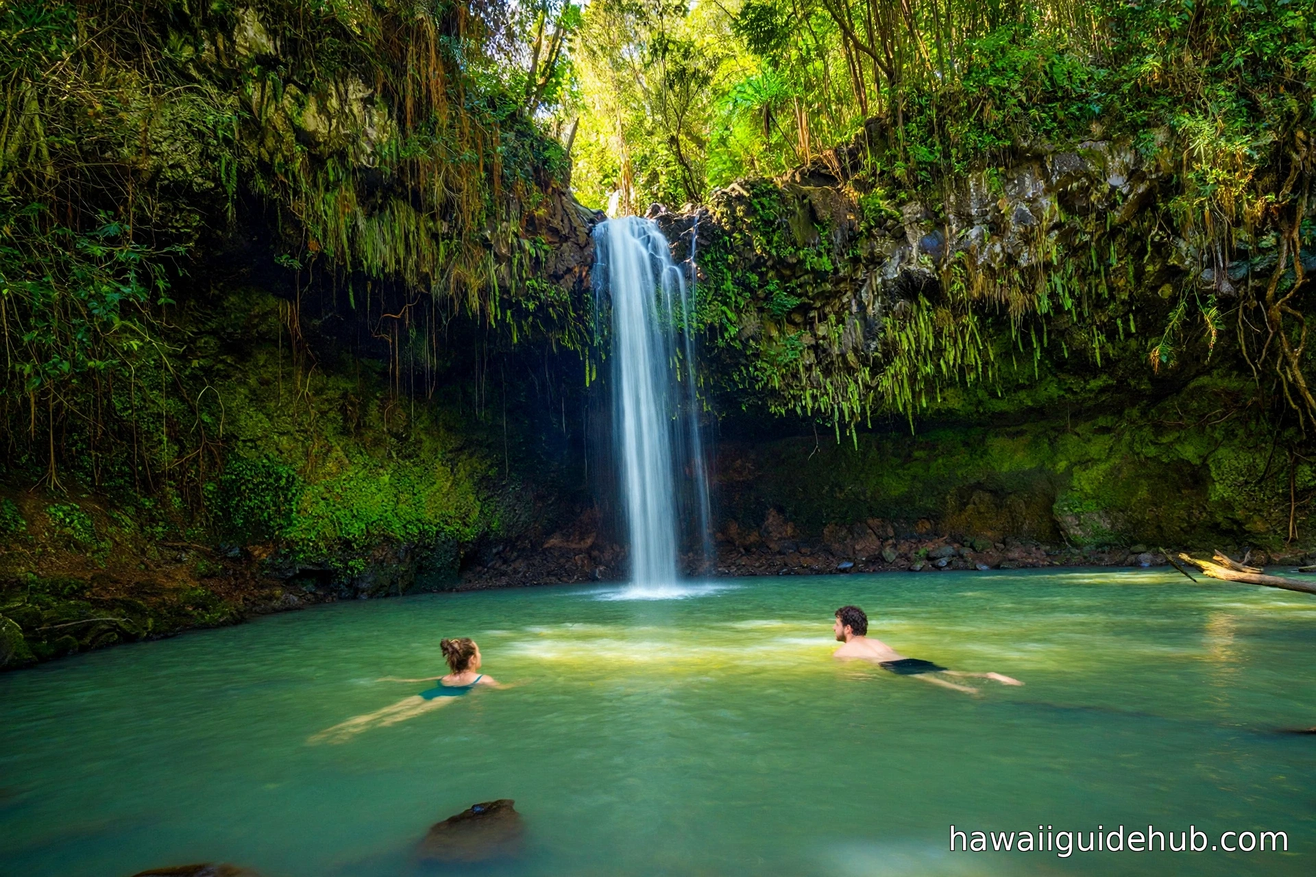 Two swimmers floating in the clear natural pool beneath a jungle waterfall at Twin Falls on the Road to Hana, Maui